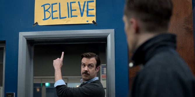 Ted Lasso pointing up to a sign that says Believe